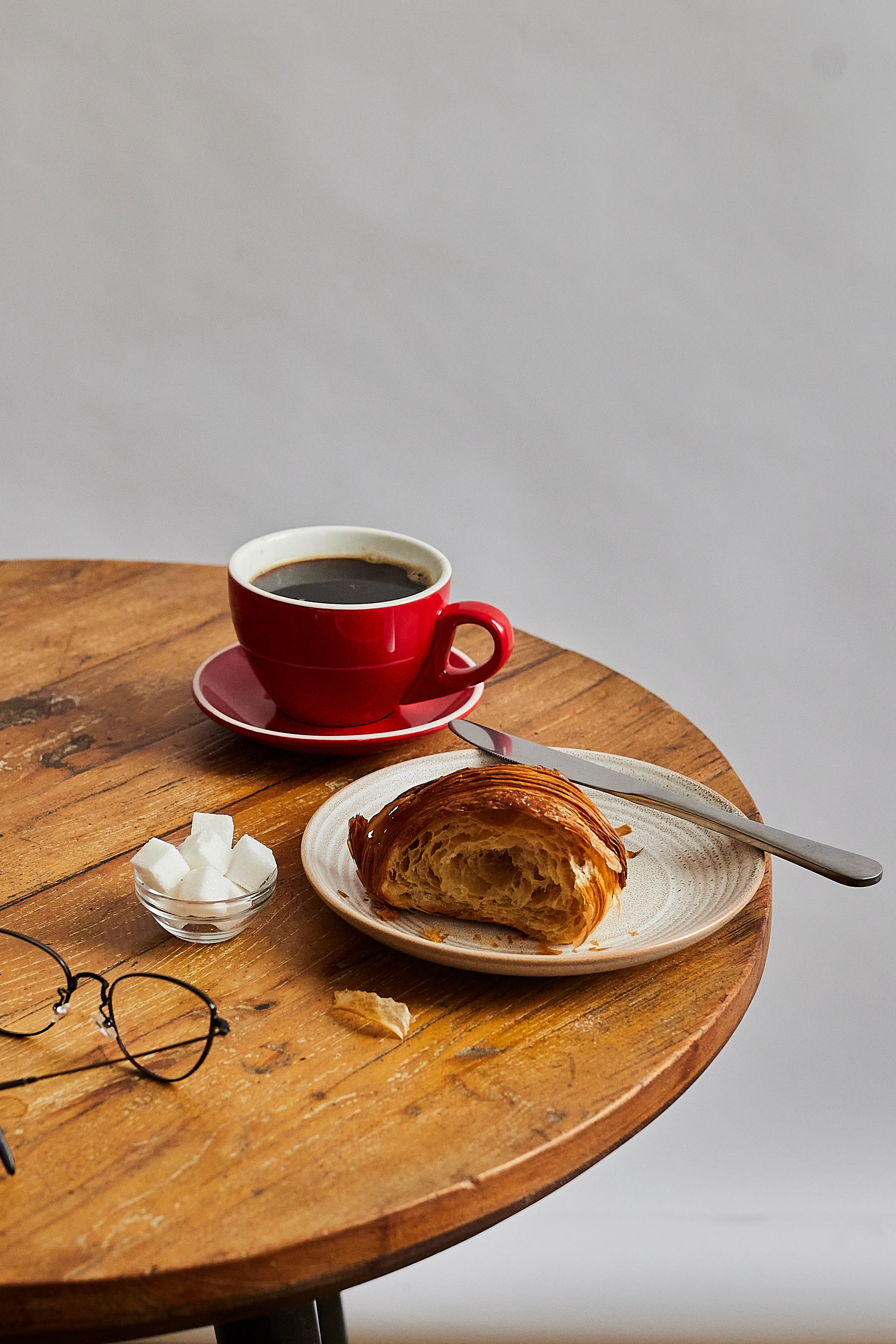 A red cup of black coffee and a half-eaten croissant on a round wooden table