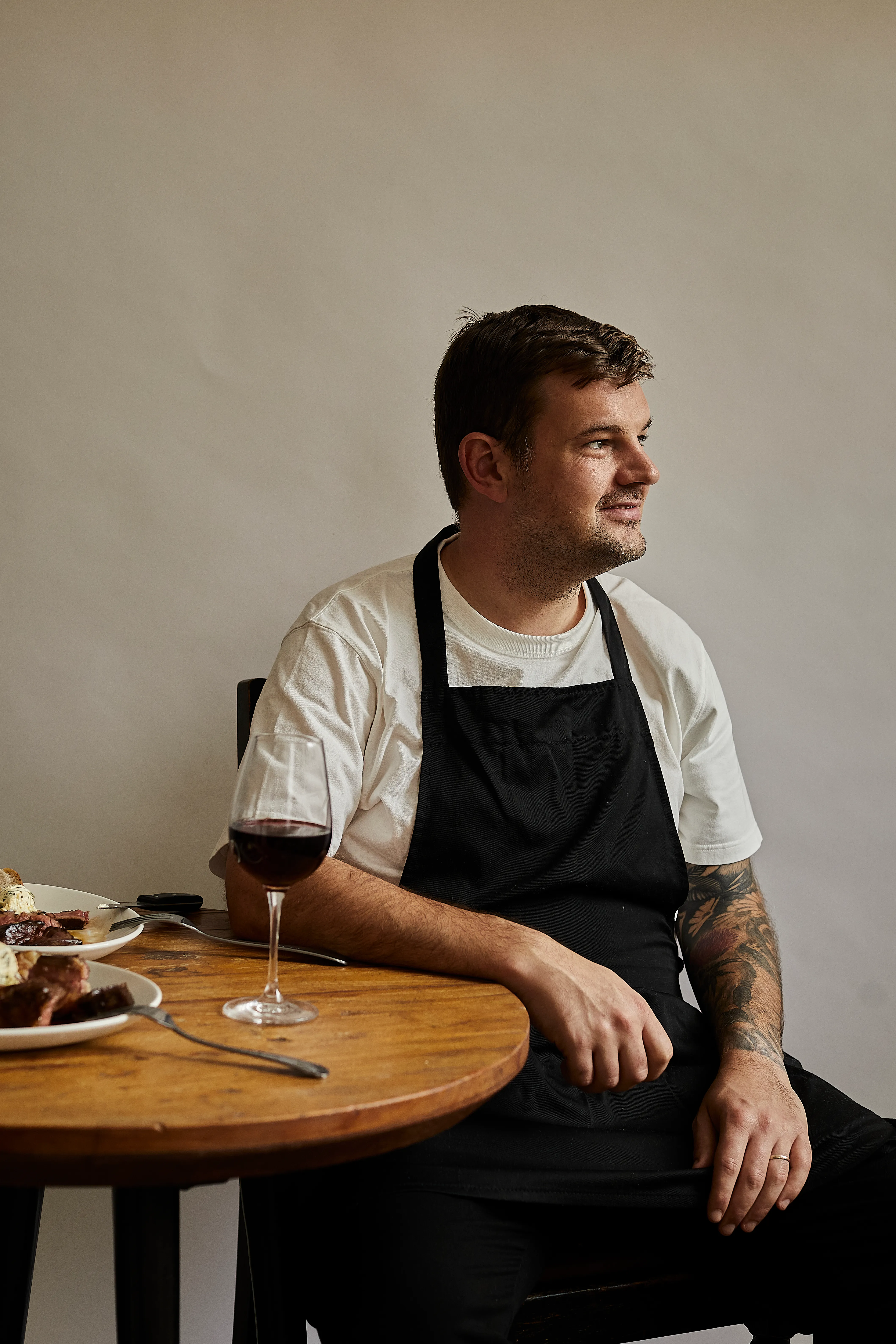 A man in an apron sitting at a wooden table with a glass of red wine, looking out of frame